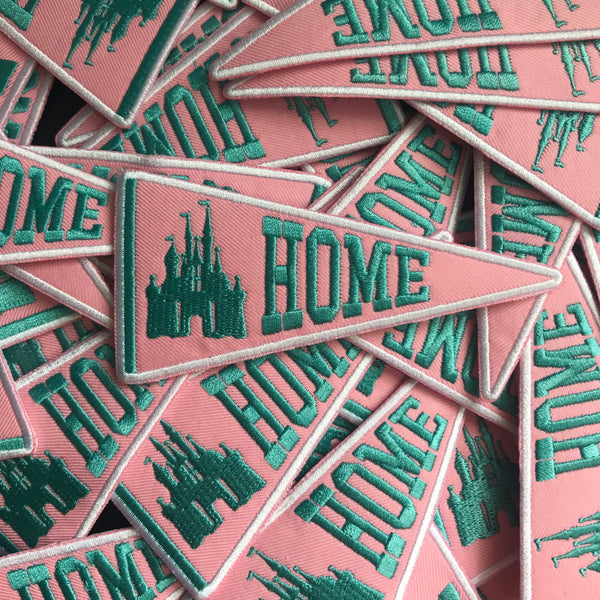 A batch of patches strewn, all of them are triangular like a banner and say HOME next to a castle in teal on top of a pink color. 