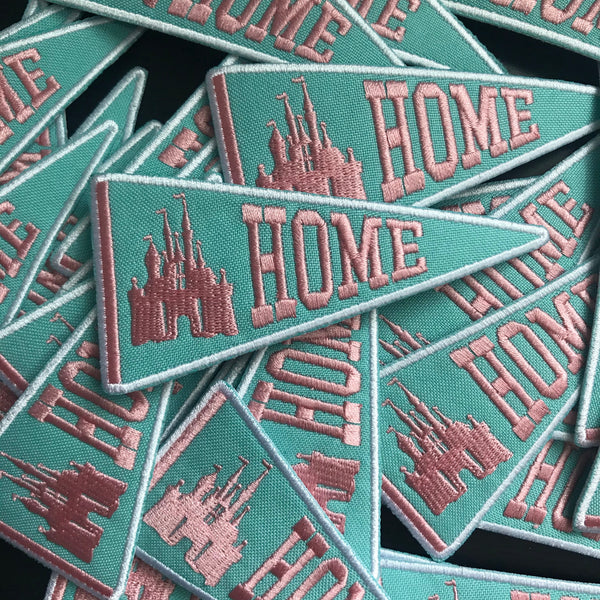 A batch of patches strewn, all of them are triangular like a banner and say HOME next to a castle in pink on top of a mint color. 